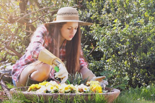 A young woman in a plaid shirt, wearing a wide-brimmed straw hat and gardening gloves, is kneeling in a lush garden, carefully planting or tending to a vibrant flower bed filled with yellow, white, and purple flowers. She is surrounded by dense green foliage, including bushes and trees, with sunlight filtering through the leaves, creating a bright and natural outdoor setting. The garden features a neatly maintained lawn with well-defined borders, and the background shows a mixture of leafy plants and shrubs, indicating a thoughtfully landscaped outdoor space typical of residential gardens in Pimlico. The scene captures a moment of peaceful gardening activity, emphasizing the care and attention to detail involved in maintaining a tidy, attractive garden area, which aligns with professional gardening services offered by Gardening Services Pimlico.